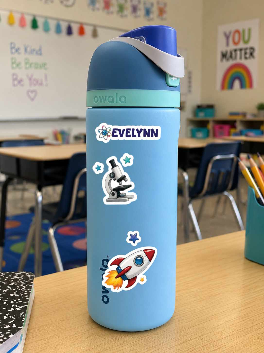 A light blue Owala water bottle with a dark blue lid and white strap, decorated with stickers including a microscope, stars, a rocket, and the name 'Evelynn', sitting on a wooden desk in a classroom.