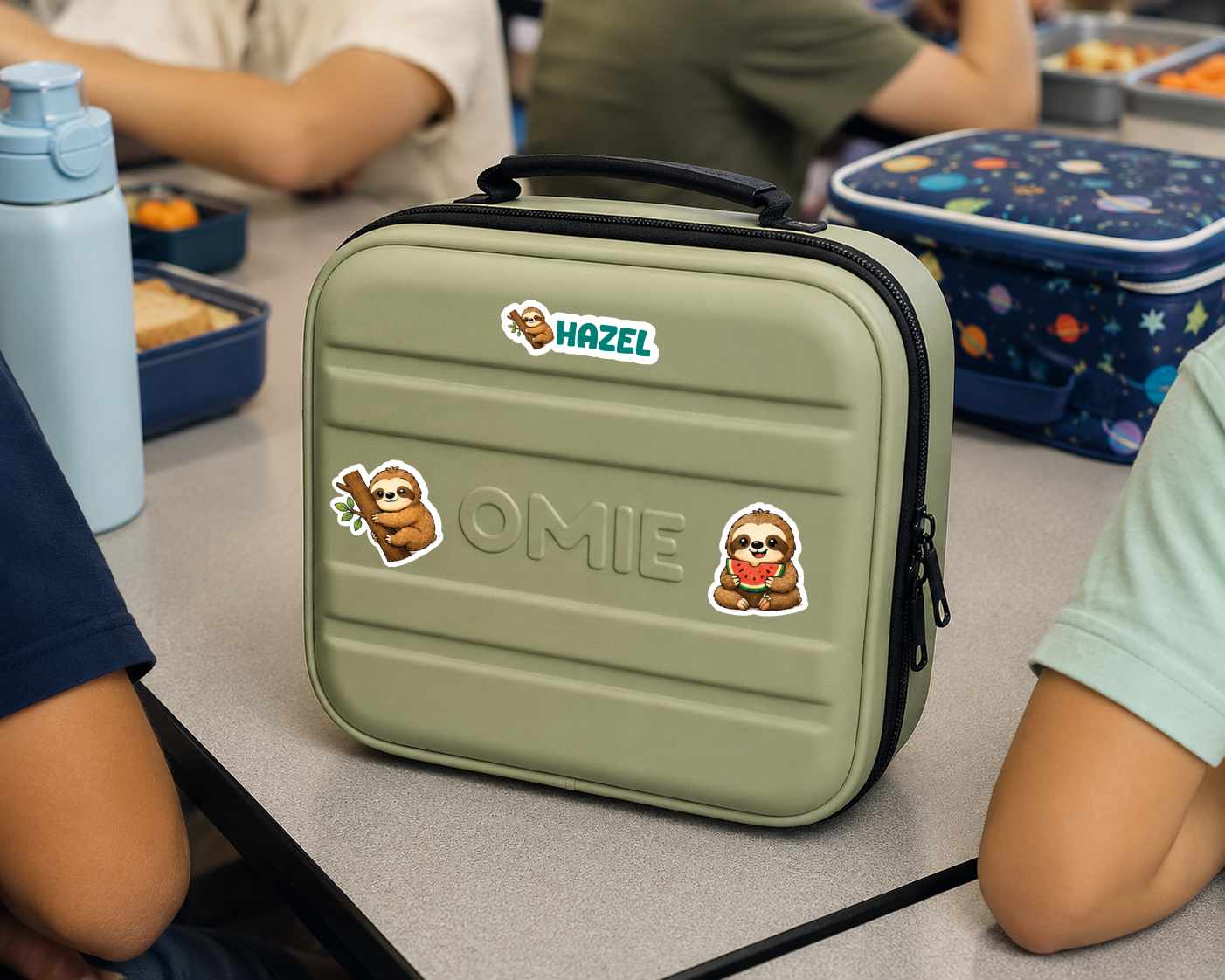 A personalized olive green hard-shell lunch bag with a black handle and zipper, decorated with stickers reading 'WILLIAM' and 'OMIE' along with cartoon characters. The bag is shown on a school desk surrounded by children and lunchboxes.
