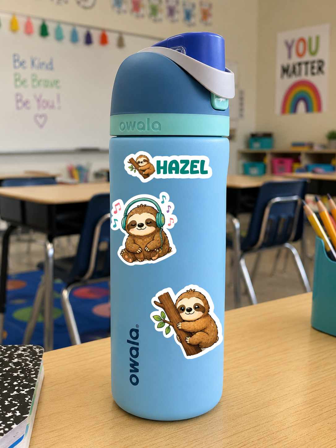 A light blue Owala water bottle with a blue lid and white strap, decorated with two sloth stickers and a 'HAZEL' name sticker, sitting on a wooden desk in a classroom.