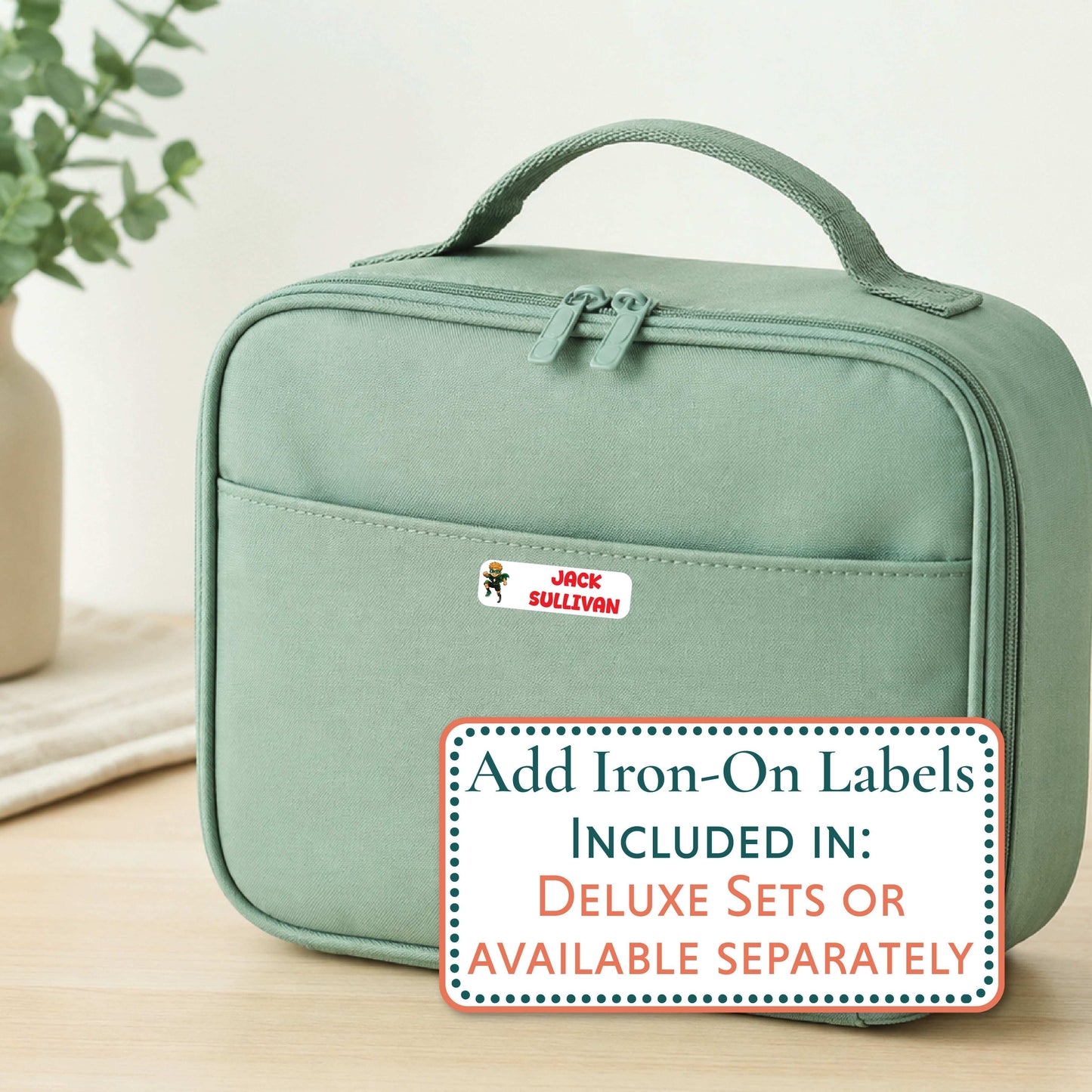A sage green insulated lunch bag with a top handle and front zip pocket, shown on a light wood surface with a decorative plant in the background. A white sticker with the name 'Aaliyah' is visible on the front pocket, and a promotional text overlay at the bottom reads 'Add Iron-On Labels INCLUDED IN: DELUXE SETS OR AVAILABLE SEPARATELY'.