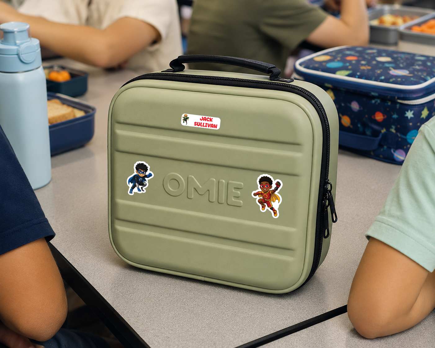 An olive green lunch bag with a black handle and zipper, decorated with stickers and the name 'OMIE' embossed on the front, sitting on a school desk with children around it.