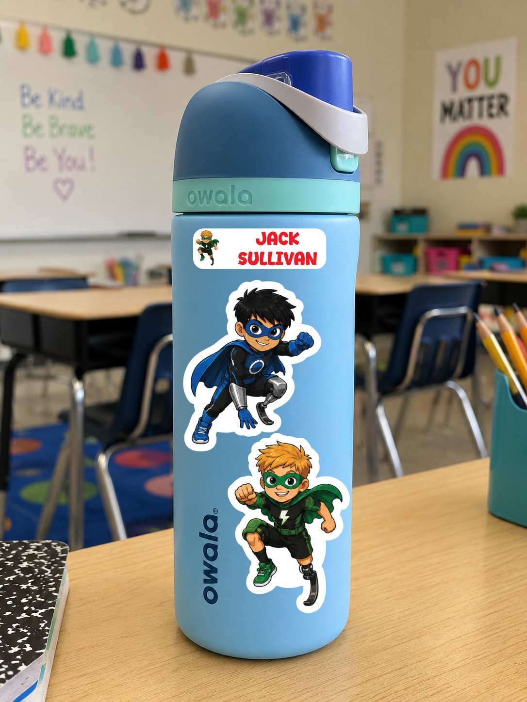 A light blue Owala water bottle with a custom name sticker and superhero character decals, standing on a wooden desk in a classroom.