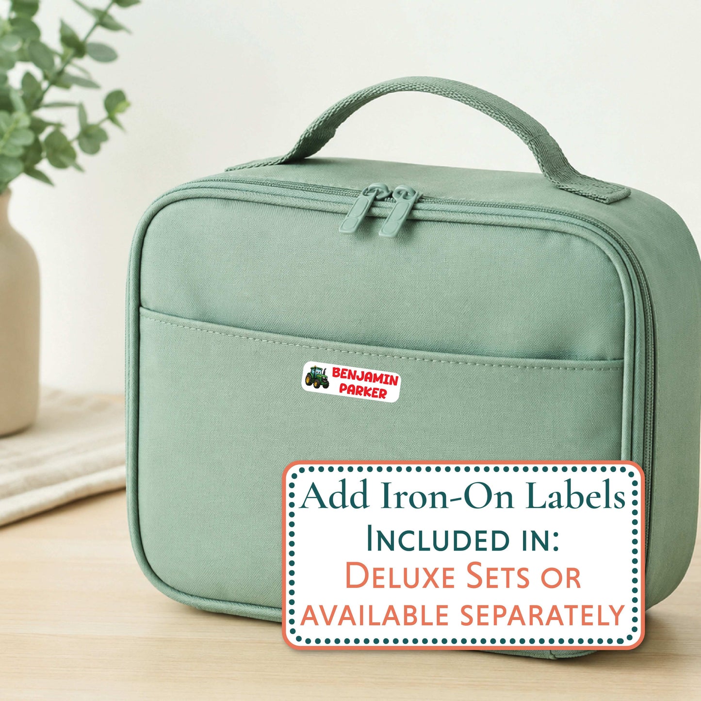 A sage green insulated lunch bag with a top handle and front zip pocket, shown on a light wood surface with a decorative plant in the background. A white sticker with the name 'Aaliyah' is visible on the front pocket, and a promotional text overlay at the bottom reads 'Add Iron-On Labels INCLUDED IN: DELUXE SETS OR AVAILABLE SEPARATELY'.