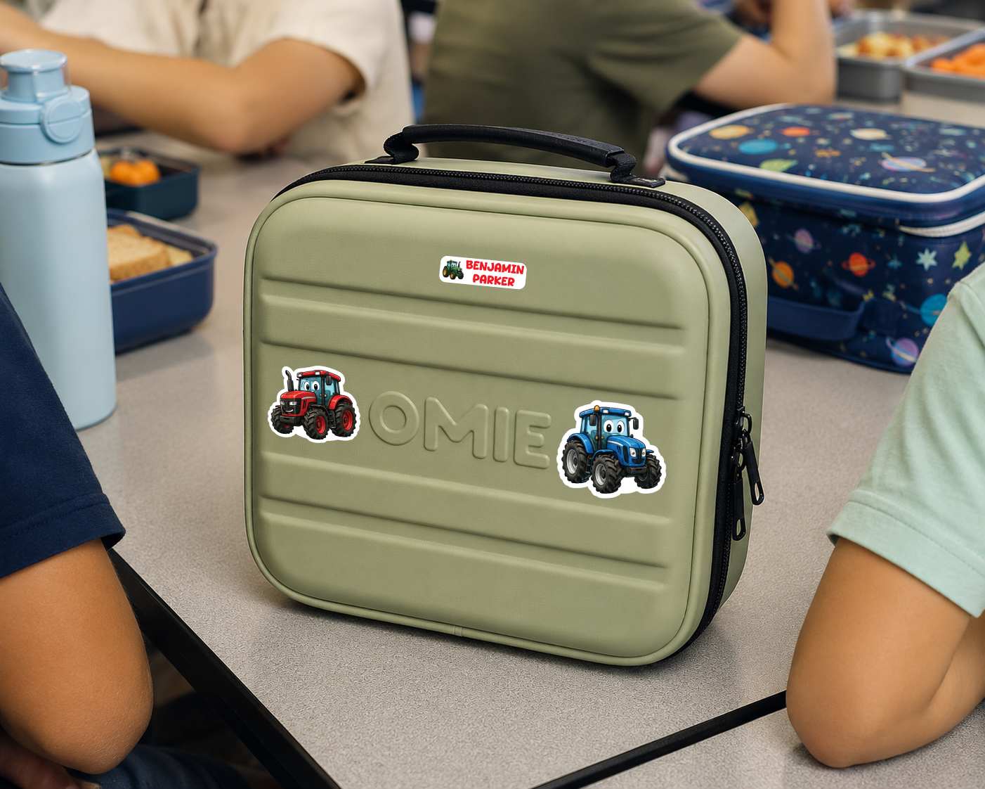 A personalized olive green hard-shell lunch bag with a black handle and zipper, featuring a custom name 'OMIE' and tractor stickers, shown on a school desk with children and lunchboxes in the background.