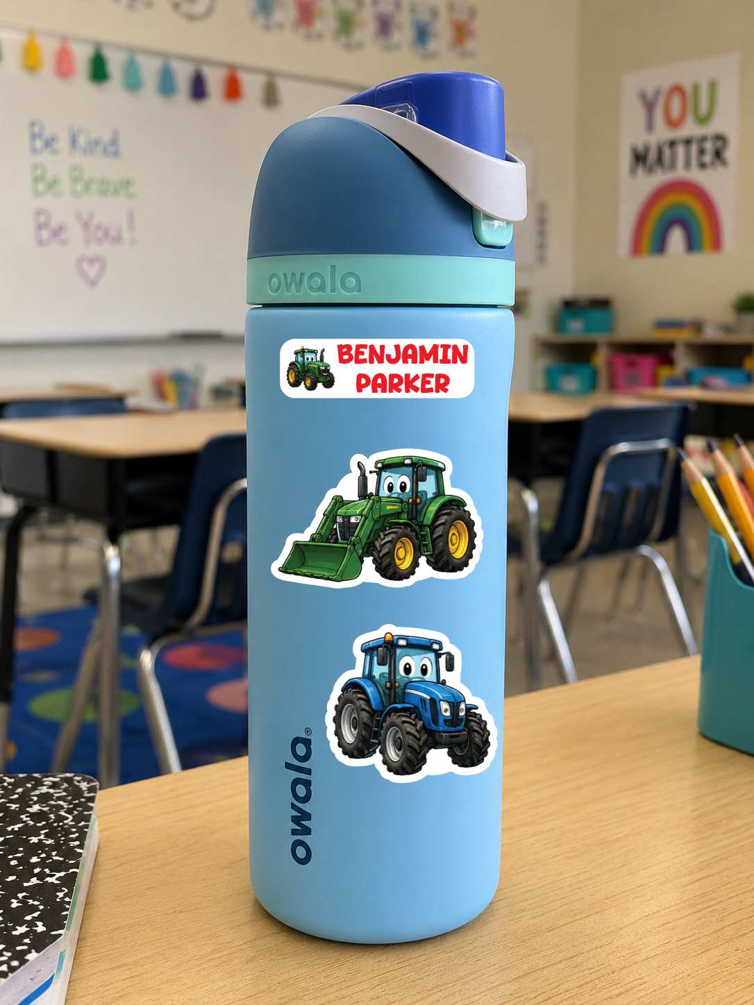 A light blue Owala water bottle with a blue lid and handle, decorated with two tractor stickers and a custom name sticker reading 'BENJAMIN PARKER', sitting on a wooden desk in a classroom.
