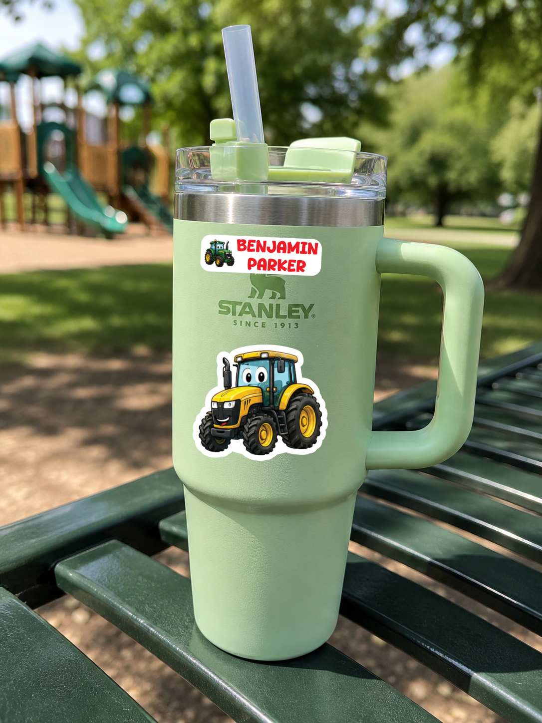 A mint green Stanley travel tumbler with a handle, clear lid, and straw, sitting on a green park bench. The tumbler features a custom sticker with the text 'BENJAMIN PARKER' and a yellow tractor illustration, with the Stanley logo printed below.