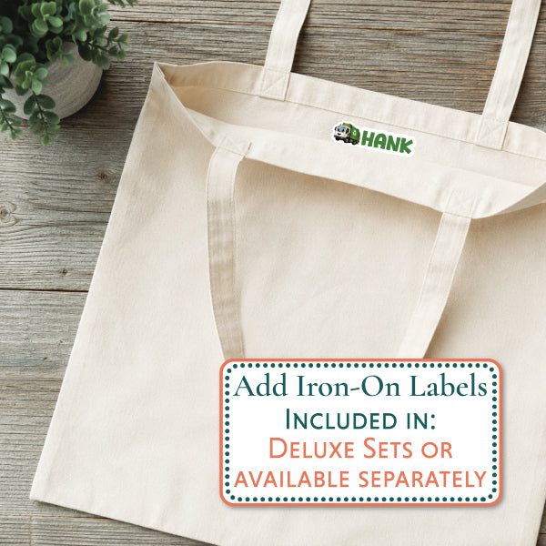 A natural canvas tote bag laid flat on a rustic wood surface, with a small green and brown 'HANK' logo printed inside the top opening. A white promotional label at the bottom reads, 'Add Iron-On Labels INCLUDED IN: DELUXE SETS OR AVAILABLE SEPARATELY.