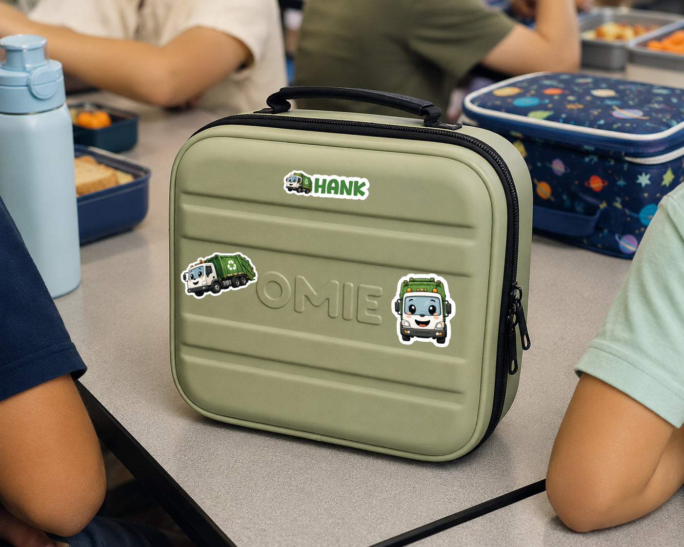 A personalized olive green lunch bag with a black handle and zipper, decorated with stickers of cartoon trash cans and the names 'HANK' and 'OMIE'. The bag is shown on a school desk surrounded by children and lunchboxes.