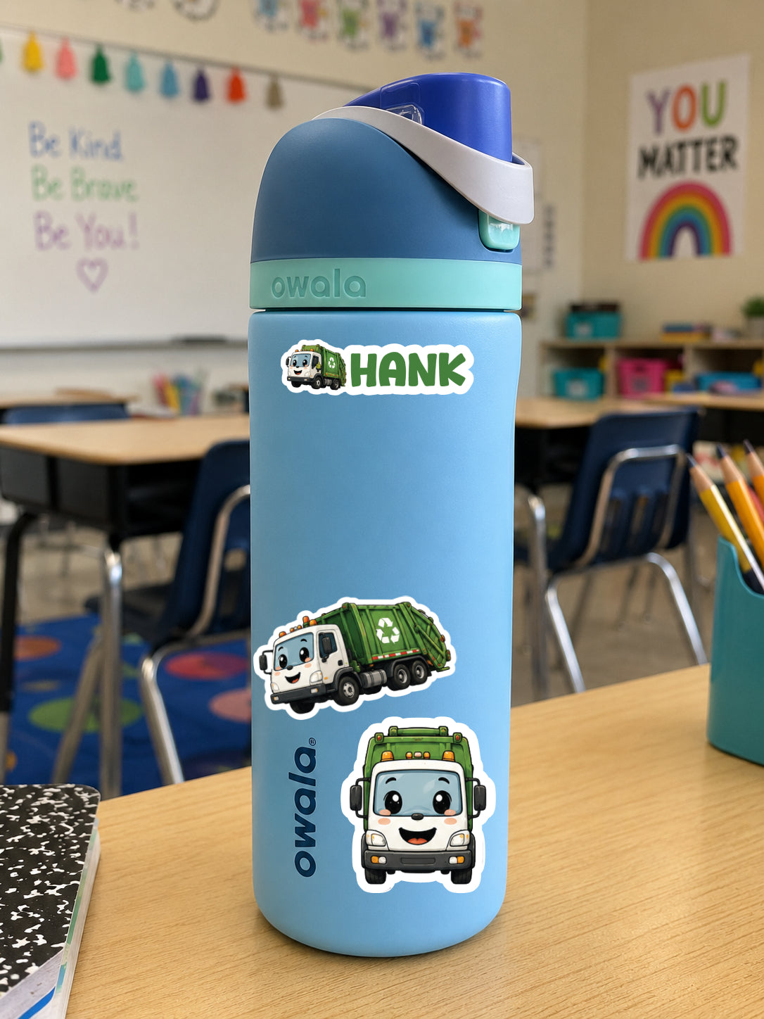 A light blue Owala water bottle with a dark blue lid and a white carrying strap, decorated with a green garbage truck sticker and the name 'HANK' on a classroom desk.
