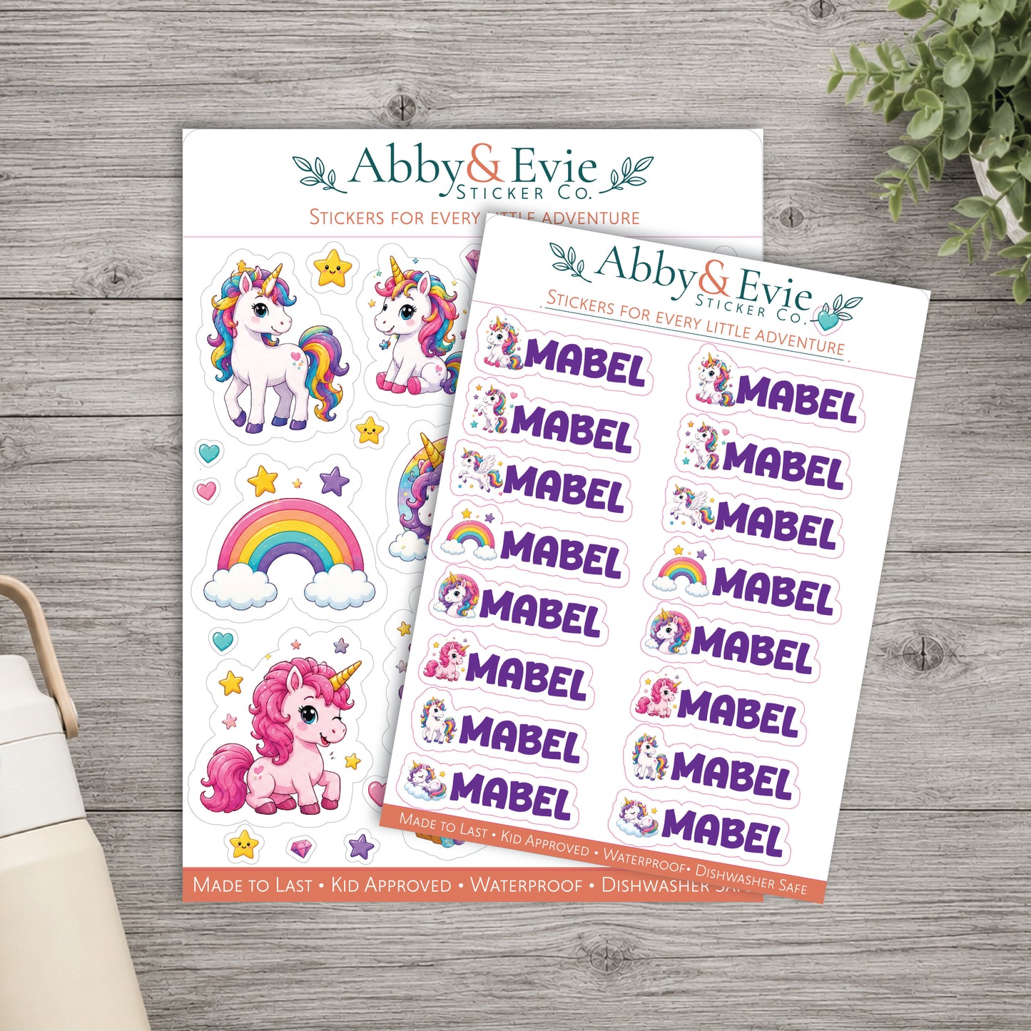Two sheets of whimsical unicorn-themed stickers from Abby & Evie Sticker Co, displayed on a gray wood background with a small potted plant and part of a bag visible.