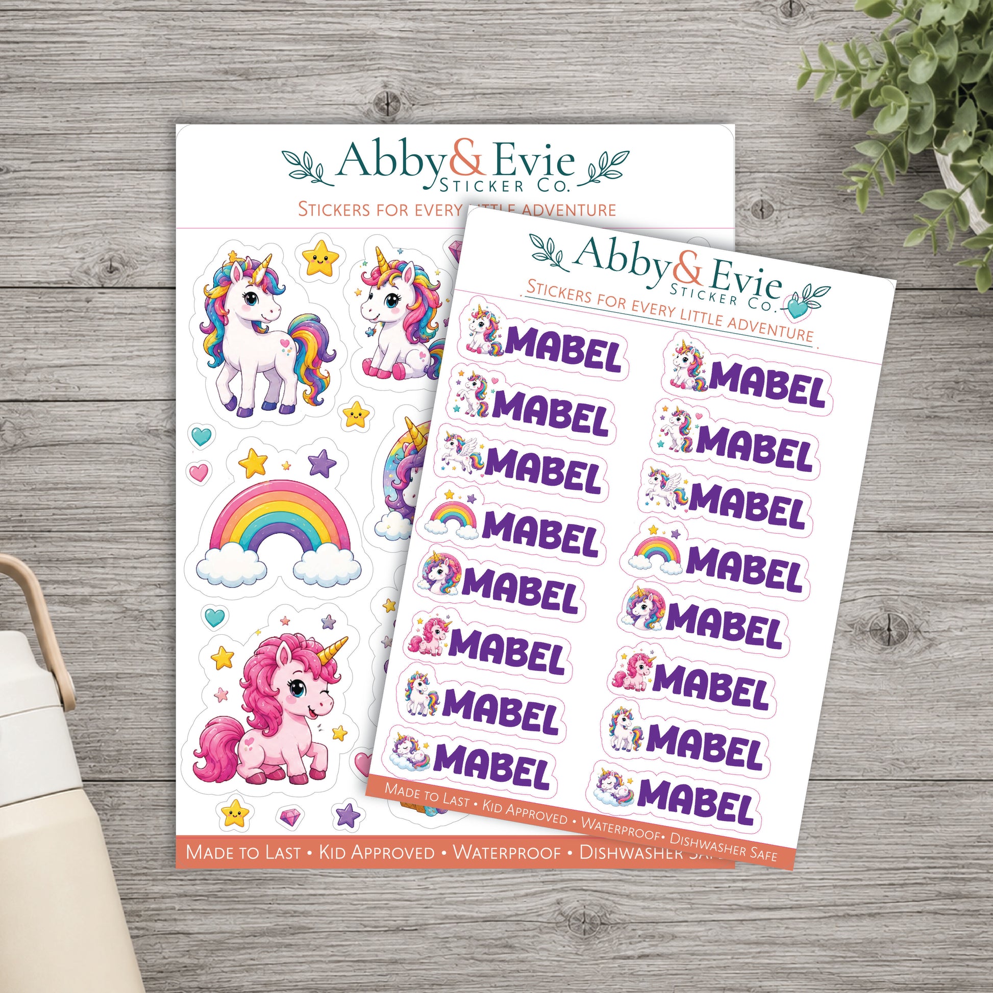 Two sheets of whimsical unicorn-themed stickers from Abby & Evie Sticker Co, displayed on a gray wood background with a small potted plant and part of a bag visible.