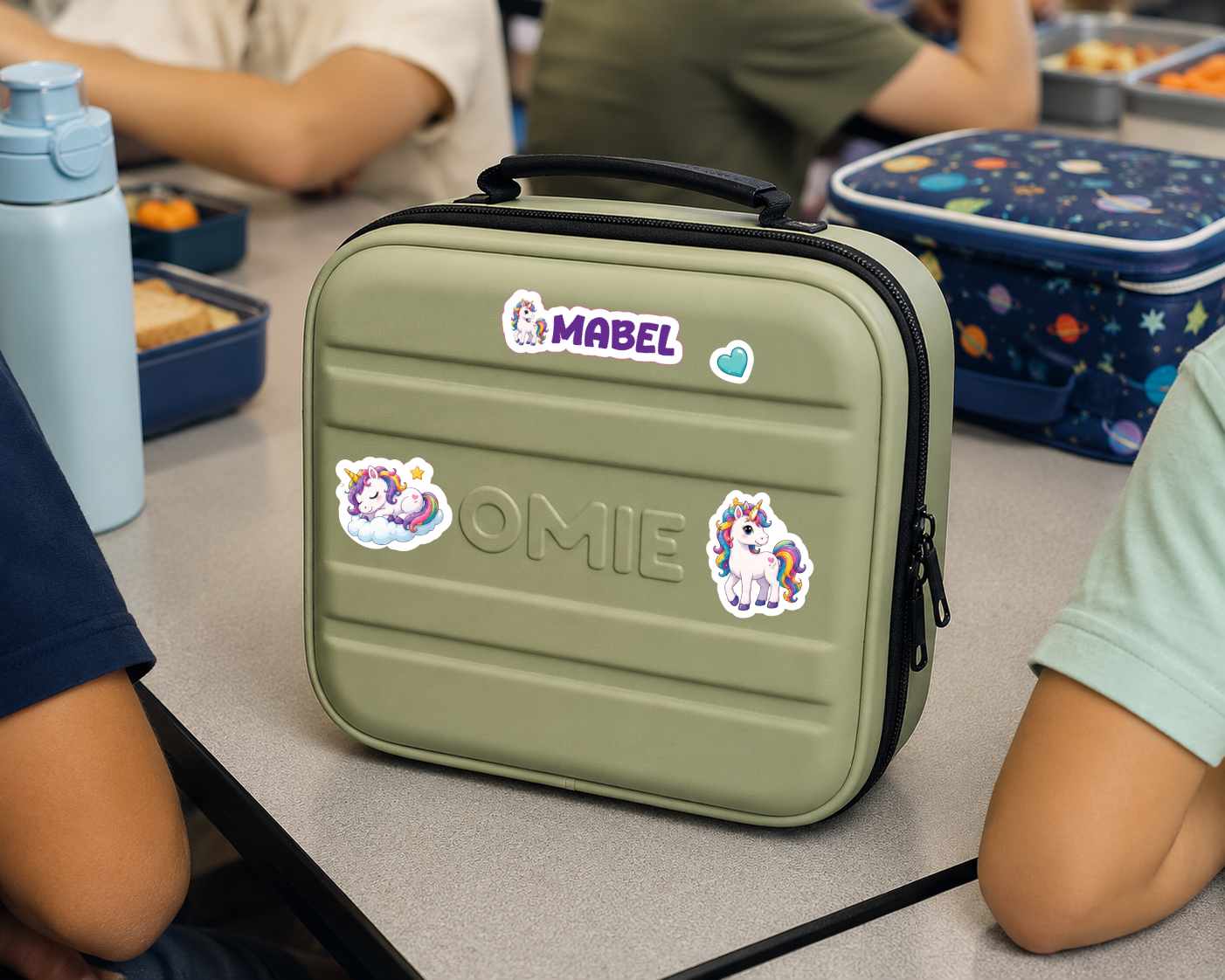 An olive green hard-shell lunch bag with a black handle and zipper, decorated with stickers including the name 'MABEL' and unicorn designs, sitting on a classroom table with children's arms and other lunchboxes visible in the background.