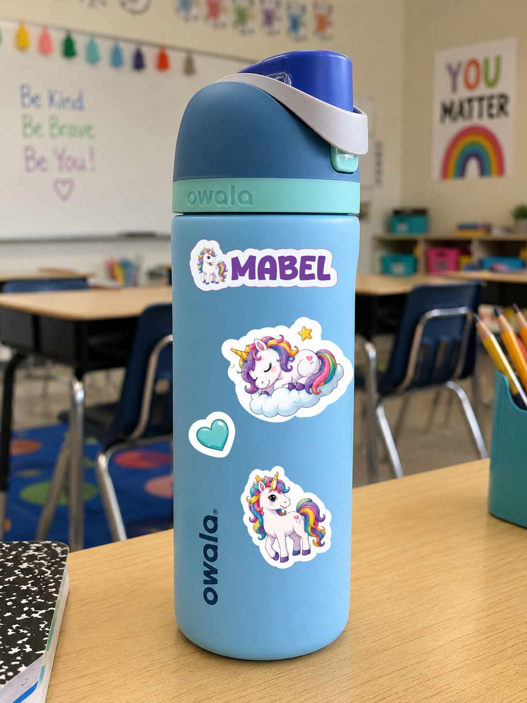 A light blue Owala water bottle with unicorn stickers, including a custom name sticker reading 'MABEL', sitting on a wooden desk in a classroom.