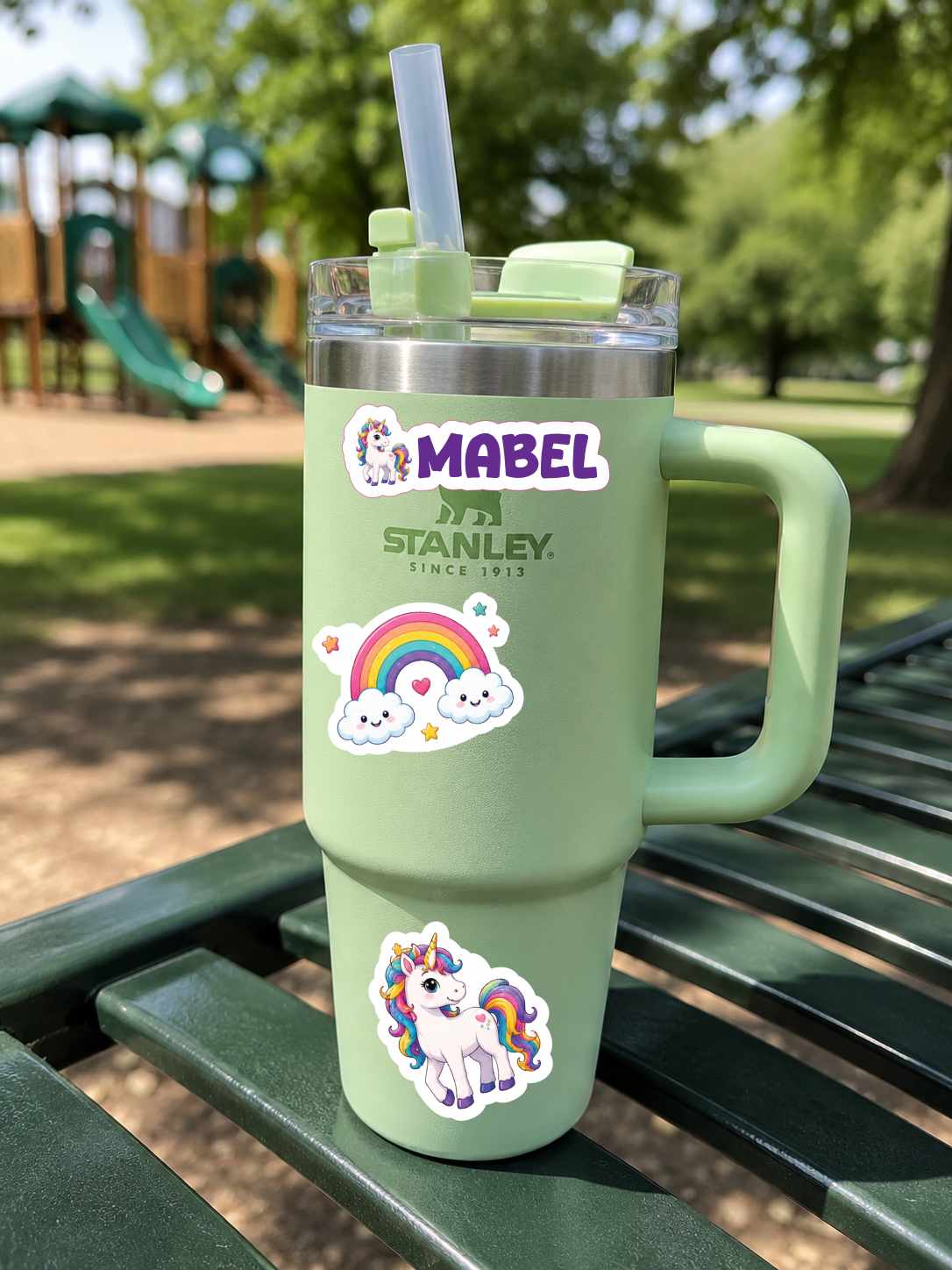 A mint green Stanley travel tumbler with a handle, clear lid, and straw, decorated with stickers including a unicorn, rainbow, and the names 'MABEL' and 'STANLEY SINCE 1917'. The tumbler is sitting on a green park bench with a blurred playground in the background.