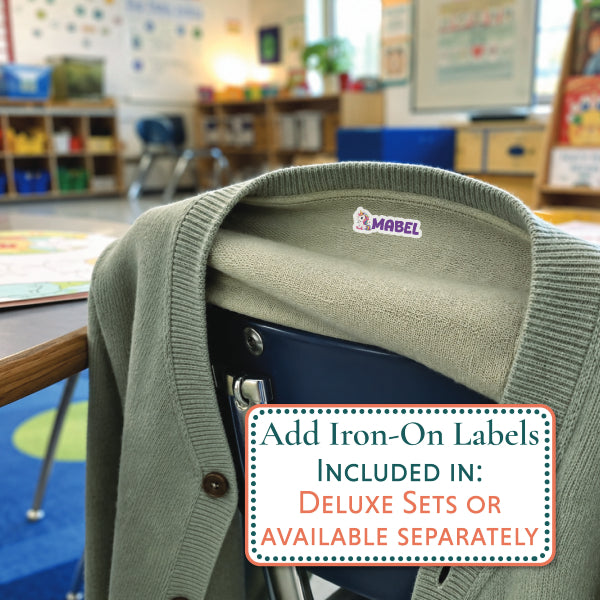 An olive green knit cardigan sweater draped over a dark chair in a blurred classroom setting, with an iron-on label sticker visible inside the collar. A text box in the foreground says, 'Add Iron-On Labels Included in: Deluxe Sets or Available Separately.