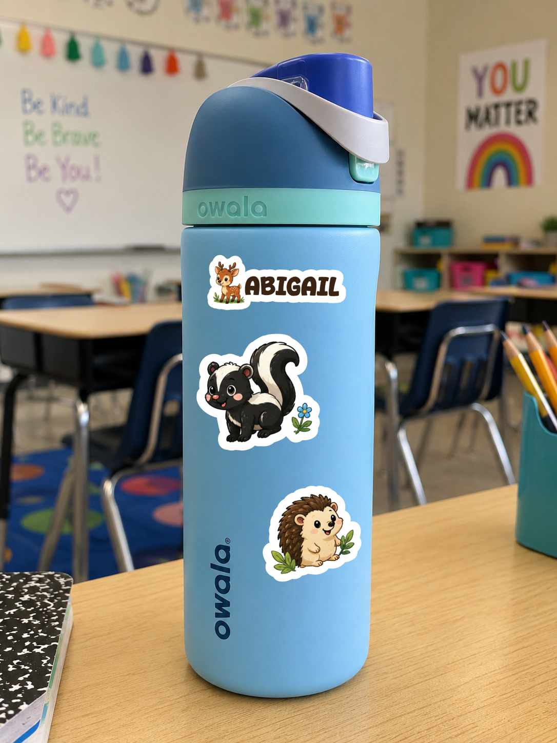 A light blue Owala water bottle with a dark blue lid and white carry loop, decorated with stickers including the name 'Abigail' and animal illustrations, sitting on a wooden desk in a classroom.