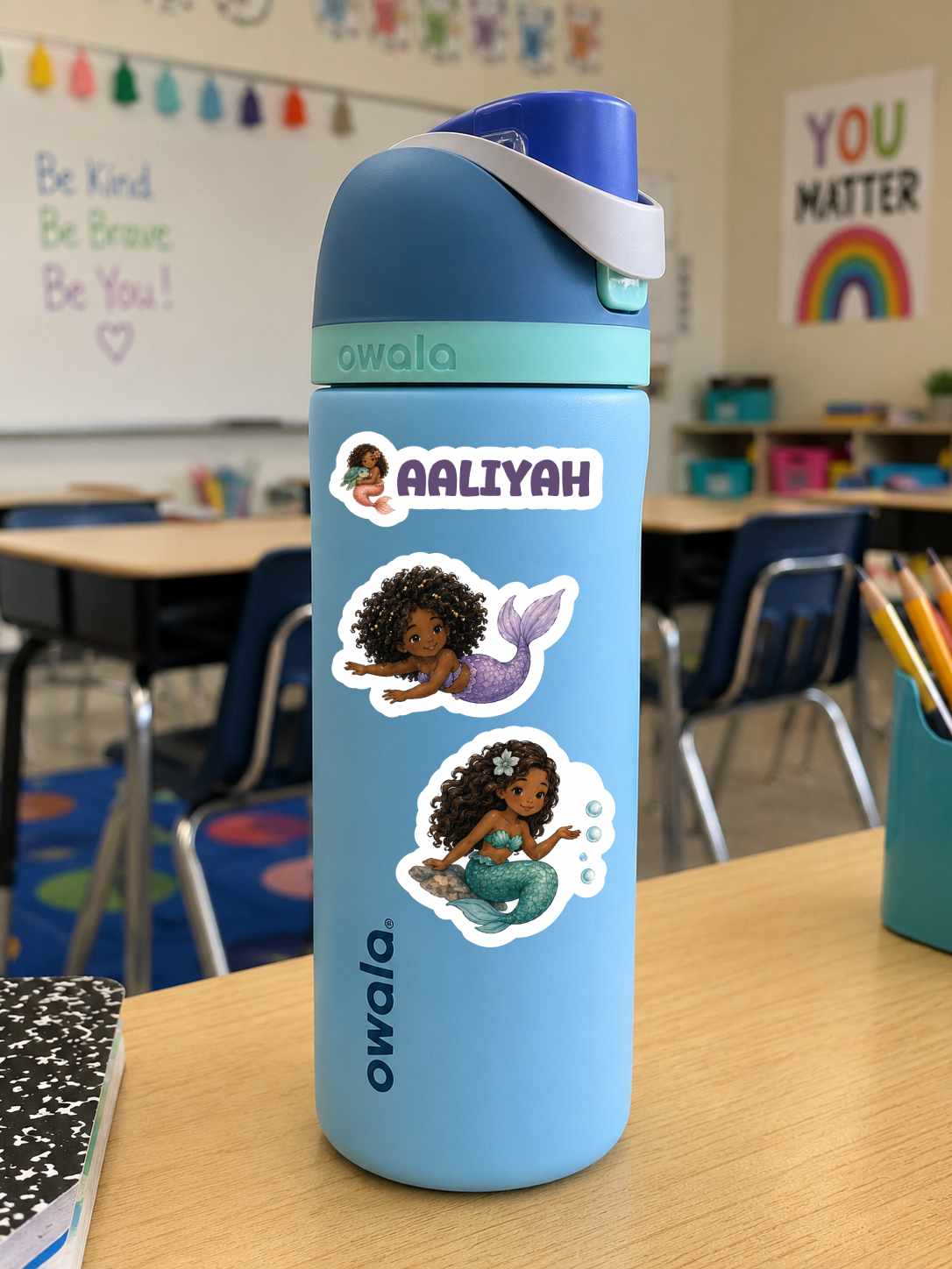 A light blue Owala water bottle with a dark blue lid and white strap, decorated with three mermaid stickers and a custom name sticker reading 'AALIIYAH', sitting on a wooden desk in a classroom.