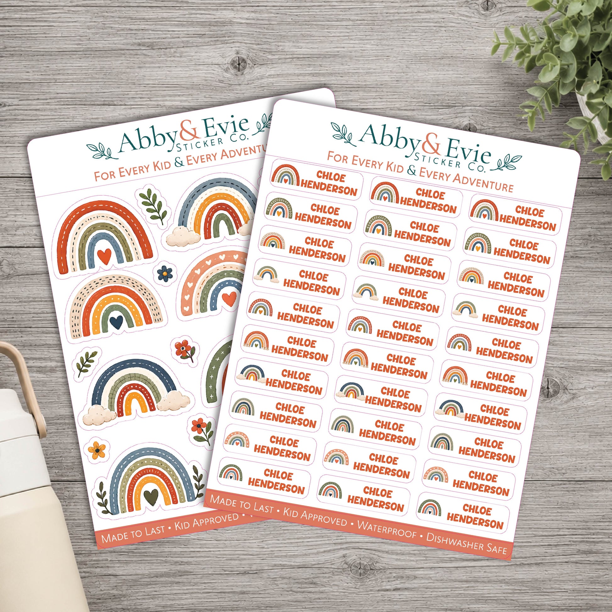 Two sticker sheets from Abby & Evie Sticker Co. featuring colorful rainbow designs and the name 'Chloe Henderson' repeated across the sheets, styled on a gray wood tabletop with a plant and notebook in the background.