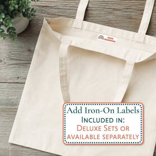 A natural canvas tote bag laid flat on a rustic wood surface, with a small purple Mabel logo patch near the top handle. A white promotional label overlay reads 'Add Iron-On Labels INCLUDED IN: DELUXE SETS OR AVAILABLE SEPARATELY.