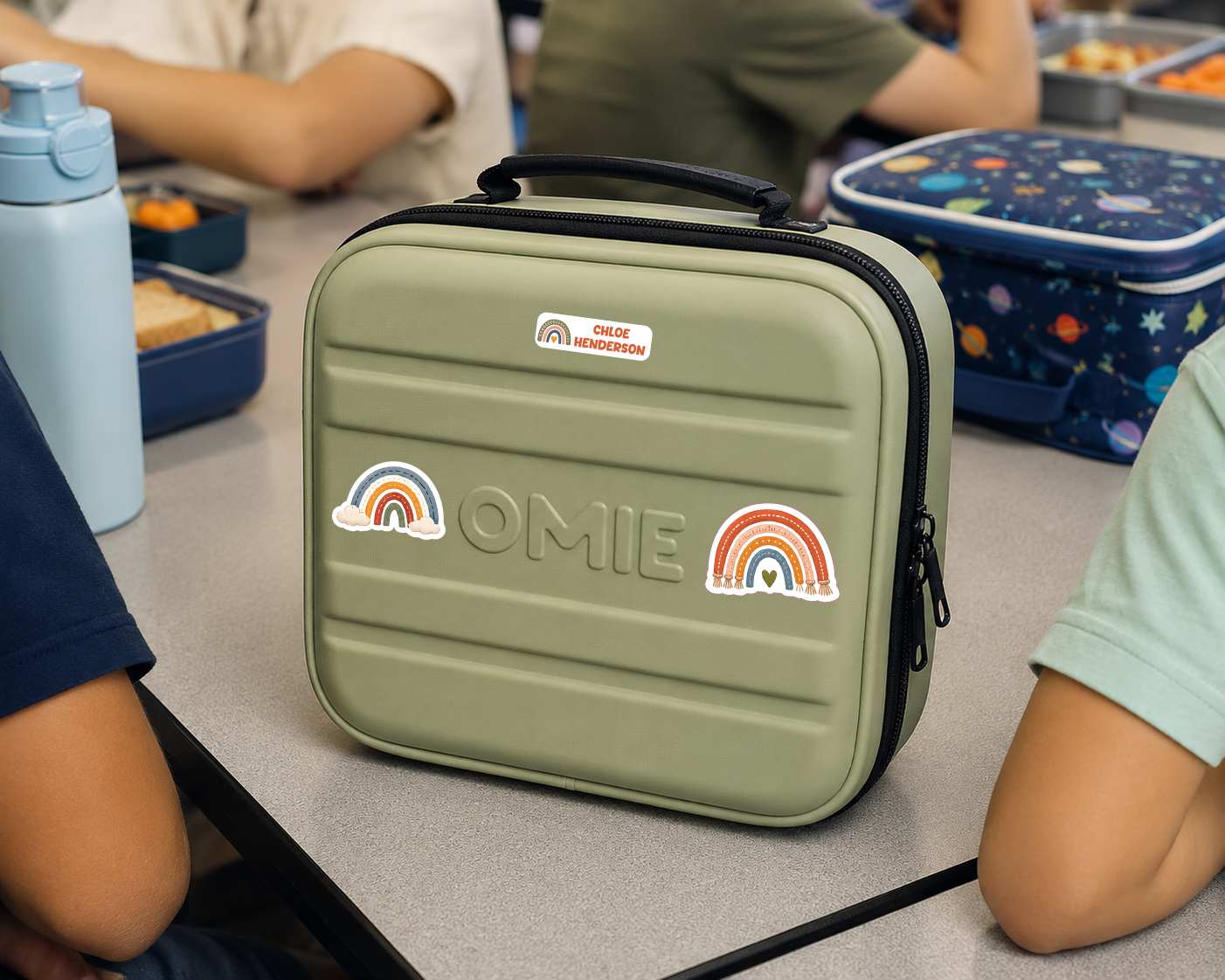 An olive green hard-shell lunch bag with a top handle, featuring the embossed text 'OMIE' and two rainbow stickers, shown on a classroom table with children seated around it.