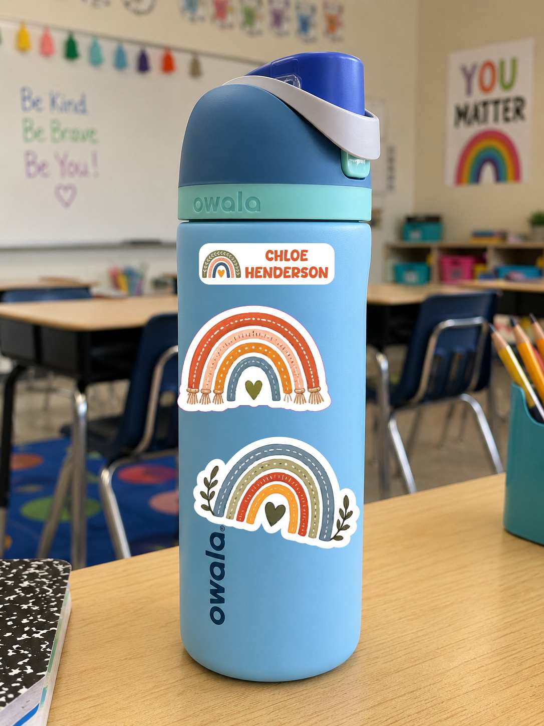 A light blue Owala water bottle with a dark blue lid and white strap, decorated with three colorful rainbow stickers, sitting on a wooden desk in a classroom.