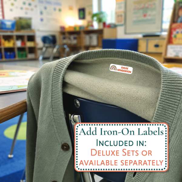 An olive green knit cardigan sweater draped over a dark chair in a blurred classroom setting. A small pink and white 'MOCKENZIE' label is visible inside the collar, and a text overlay at the bottom reads, 'Add Iron-On Labels INCLUDED IN: DELUXE SETS OR AVAILABLE SEPARATELY'.