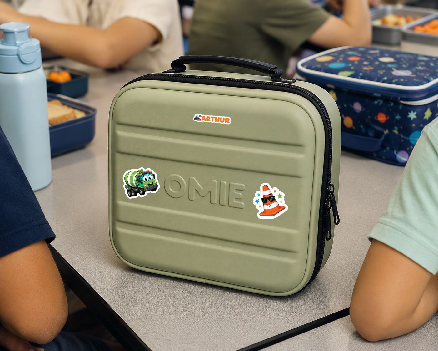 An olive green lunch bag with a black handle and zipper, decorated with stickers and the name 'OMIE' embossed on the front, sitting on a school desk with children around it.