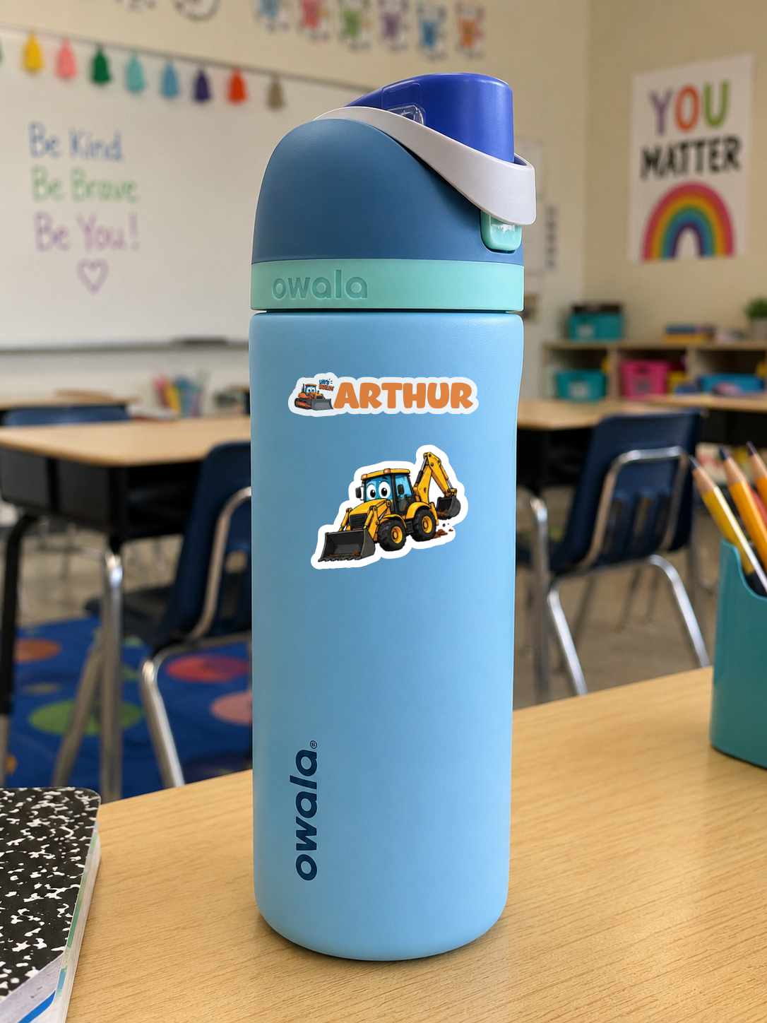 A light blue Owala water bottle with a custom 'Arthur' construction worker sticker, sitting on a wooden desk in a classroom setting.