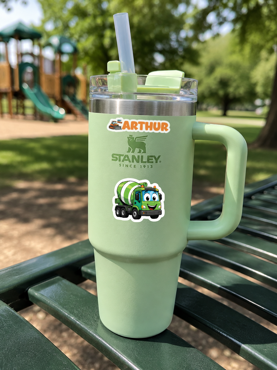 A mint green Stanley Arthur tumbler with a handle, clear lid, and straw, sitting on a green park bench with a playground in the background. The tumbler features the Arthur Stanley logo and a sticker of a green cartoon cement mixer truck.