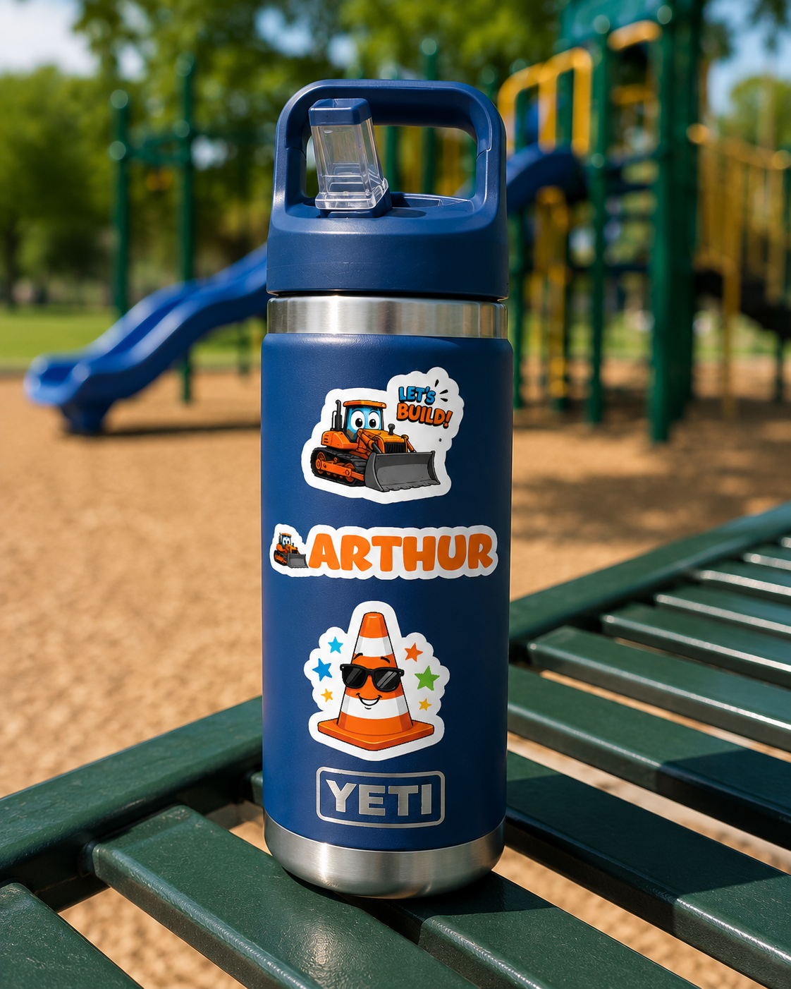 A blue YETI water bottle with custom cartoon stickers sits on a green playground bench with a blurred playground in the background.