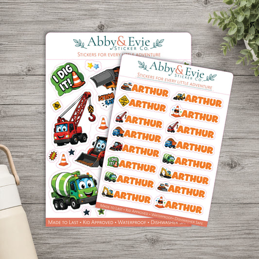 Two sheets of custom children's stickers from Abby & Evie Sticker Co. displayed on a gray wood tabletop with a small plant and part of a mug visible as styled props.