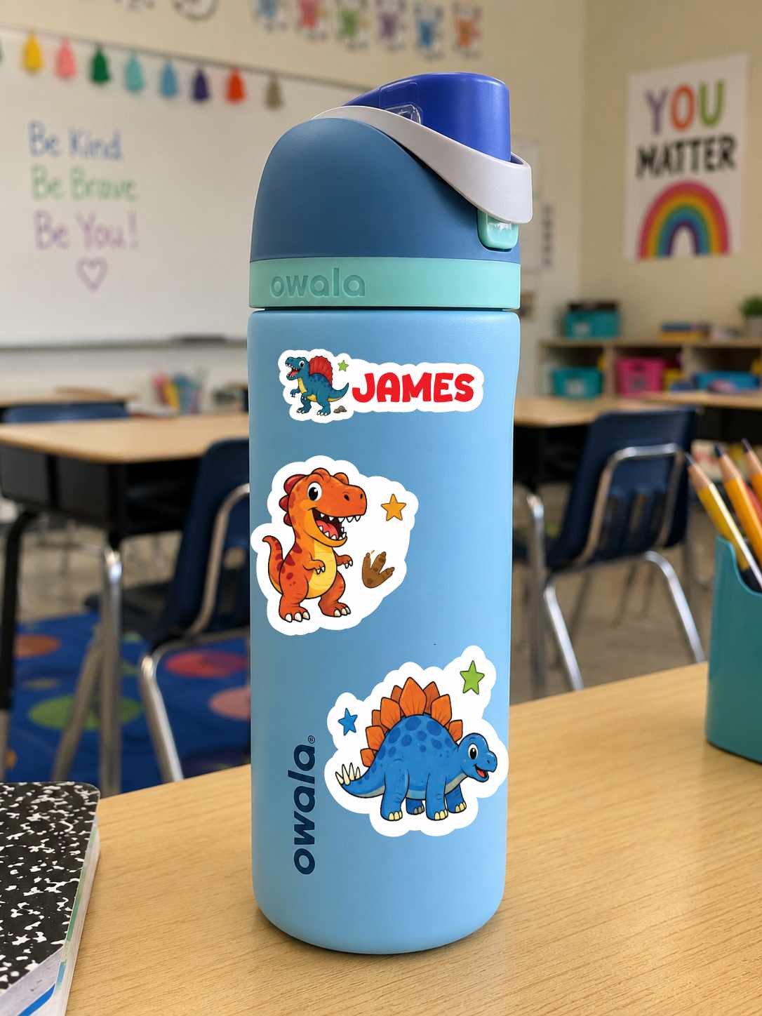 A light blue Owala water bottle with dinosaur stickers and a custom name decal reading 'JAMES', standing on a wooden desk in a classroom.