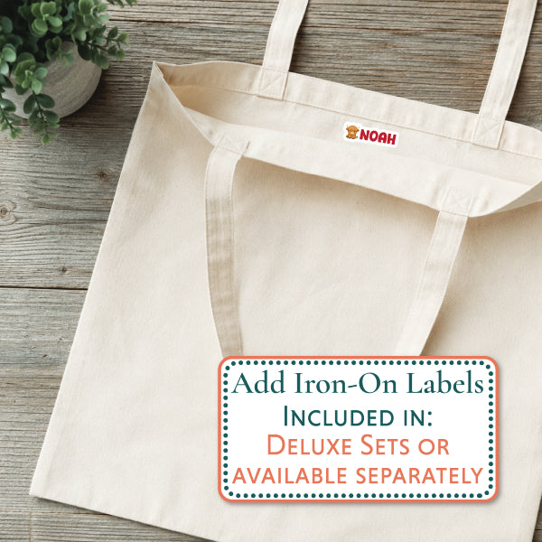 A natural canvas tote bag laid flat on a rustic wood surface, with a small red and yellow NOAH logo printed inside the neck. A white rectangular overlay at the bottom right reads, 'Add Iron-On Labels INCLUDED IN: DELUXE SETS OR AVAILABLE SEPARATELY.