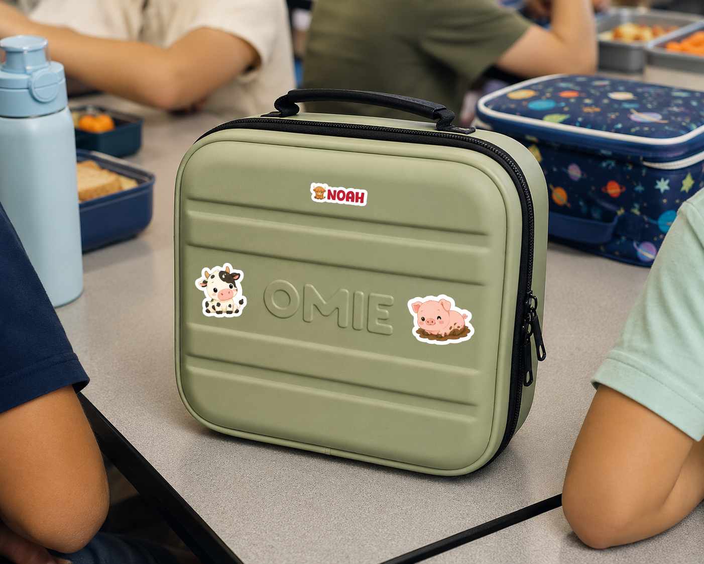 An olive green hard-shell lunch bag with a top handle, featuring the brand name NORH and cute animal stickers, sitting on a school desk with children around it.