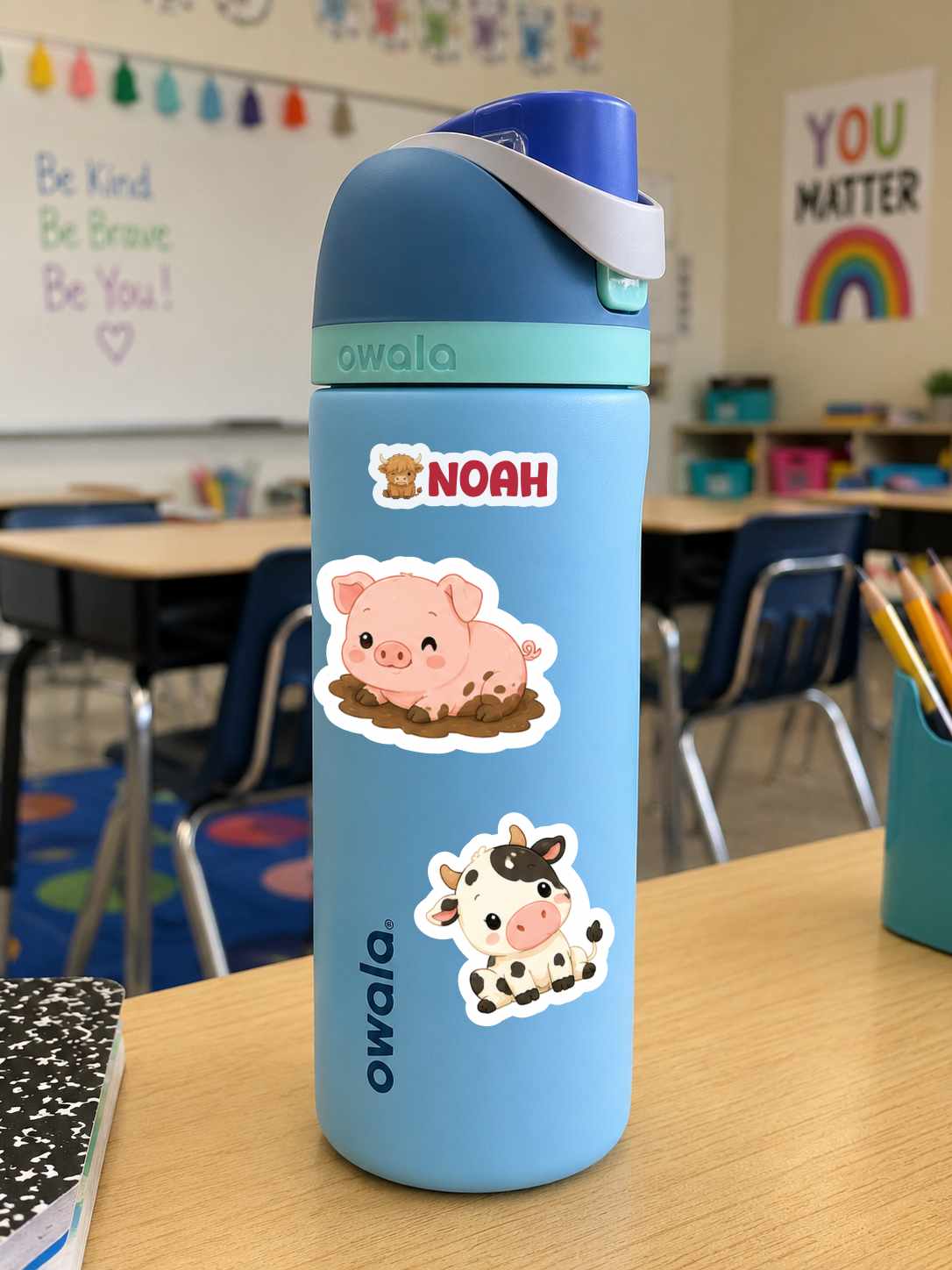 A light blue Owala water bottle with a dark blue lid and strap, decorated with two cartoon animal stickers (a pig and a cow), sitting on a wooden desk in a classroom.