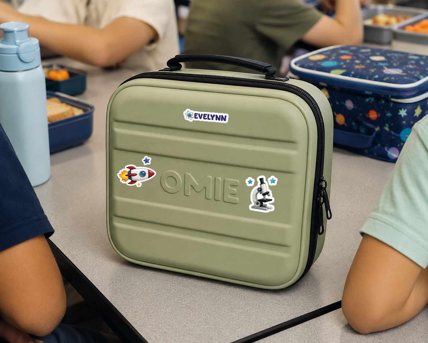 An olive green hard-shell lunch bag with a black handle and zipper, decorated with stickers and the name 'OMIE' on the front. It is shown on a classroom table surrounded by children and lunchboxes.