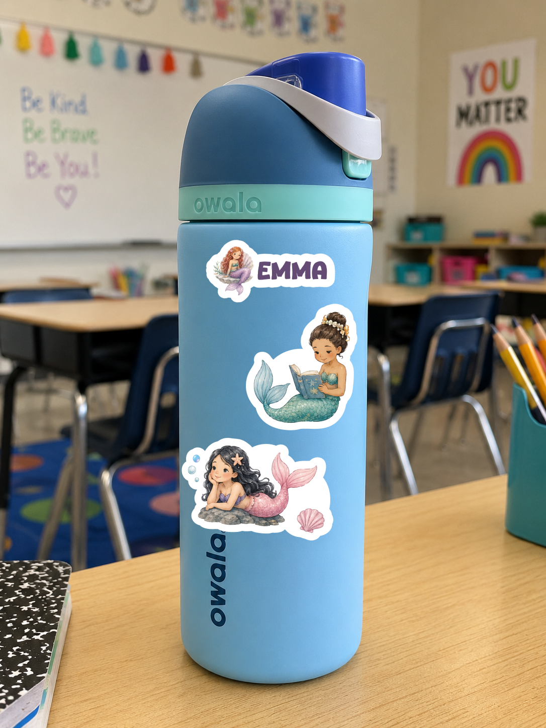 A light blue Owala water bottle with a dark blue lid and a white carrying strap, decorated with three mermaid stickers and the name 'EMMA' on a classroom desk.
