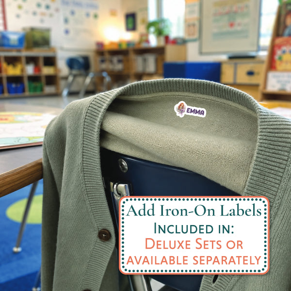 An olive green knit cardigan draped over a chair in a classroom setting, with an iron-on label sticker reading 'ARTHUR' applied to the collar. A text overlay at the bottom right says, 'Add Iron-On Labels INCLUDED IN: DELUXE SETS OR AVAILABLE SEPARATELY'.