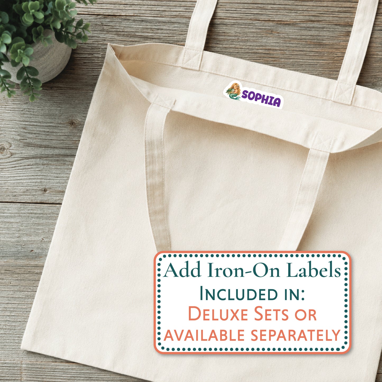 A natural canvas tote bag laid flat on a rustic wood surface, with a small purple Mabel logo patch near the top handle. A white promotional label overlay reads 'Add Iron-On Labels INCLUDED IN: DELUXE SETS OR AVAILABLE SEPARATELY.