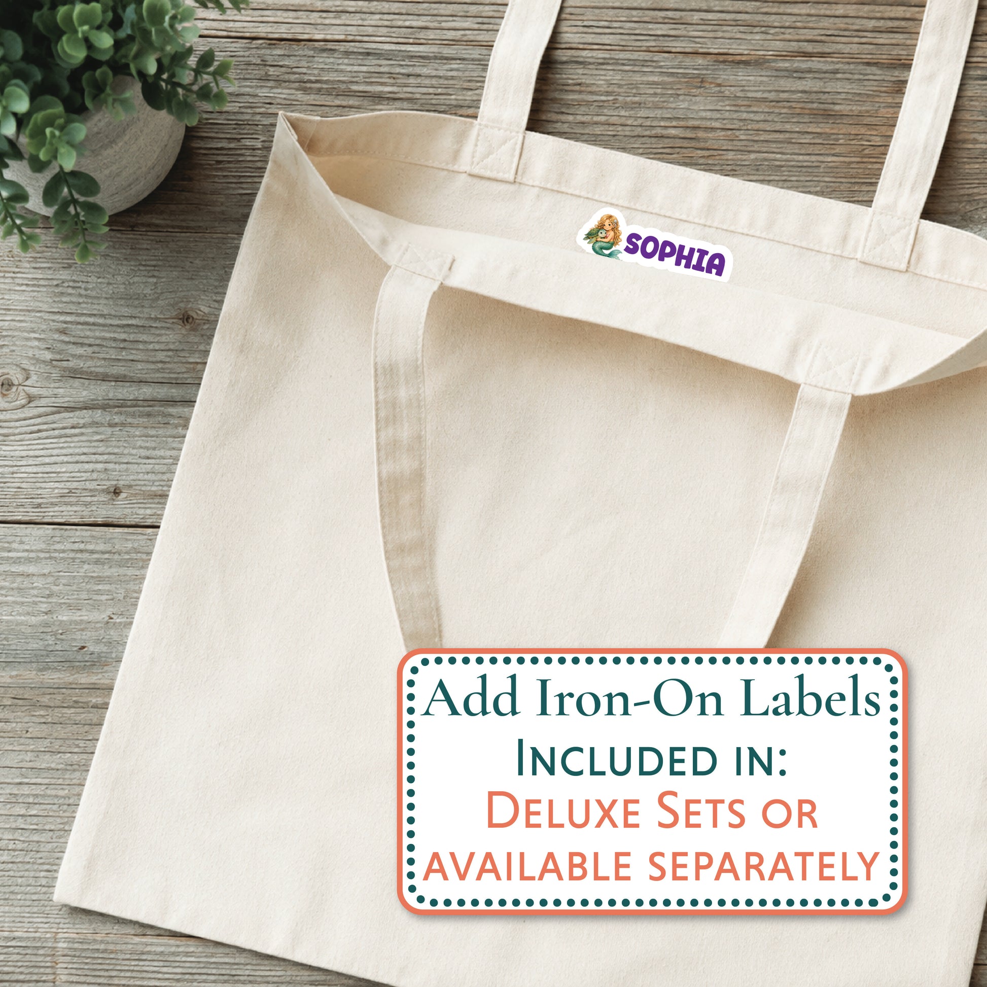 A natural canvas tote bag laid flat on a rustic wood surface, with a small purple Mabel logo patch near the top handle. A white promotional label overlay reads 'Add Iron-On Labels INCLUDED IN: DELUXE SETS OR AVAILABLE SEPARATELY.