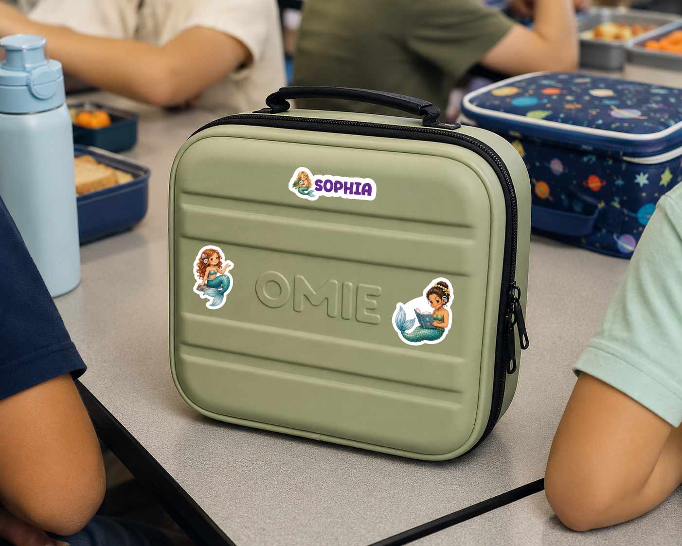 A personalized olive green hard-shell lunch bag with a black handle and zipper, decorated with stickers reading 'SOPHIA' and 'OMIE' along with cartoon mermaids with cochlear implants  illustrations, shown on a school desk with children seated around it.