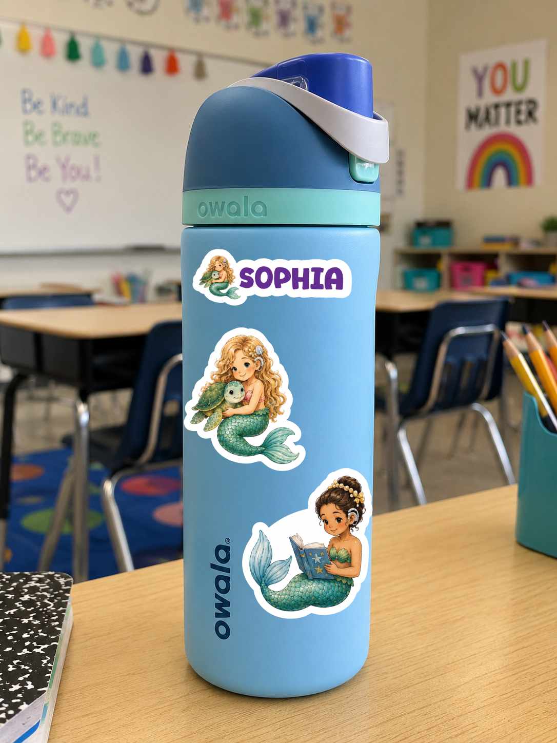 A light blue Owala water bottle with a dark blue lid and white strap, decorated with three mermaids with cochlear implants stickers and the name 'SOPHIA' in purple. The bottle is sitting on a wooden desk in a classroom setting with desks and chalkboard visible in the background.