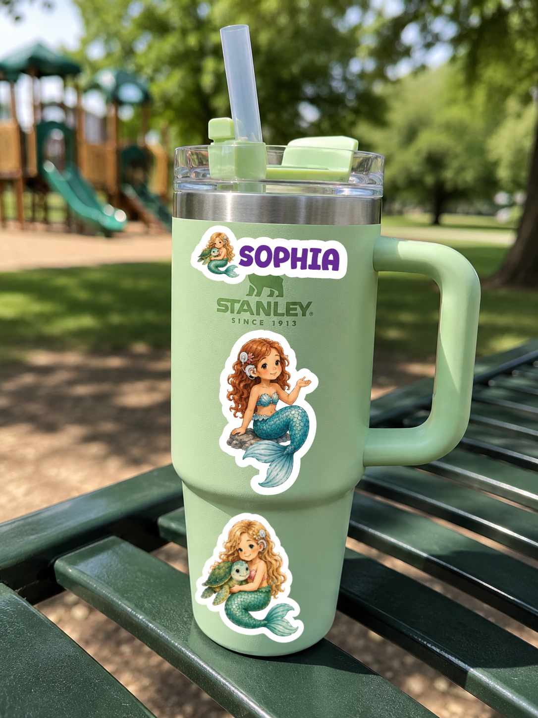 A mint green handled tumbler with a clear lid and straw sits on a green park bench. The tumbler has custom stickers featuring mermaids and the text 'SOPHIA' and 'STANLEY SINCE 1817'.
