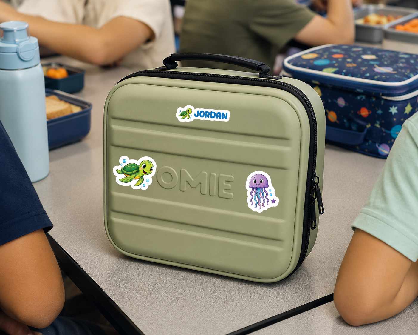 An olive green Jordan lunch bag with custom name 'OMIE' and cartoon stickers, sitting on a school desk with children's lunchboxes and arms visible in the background.
