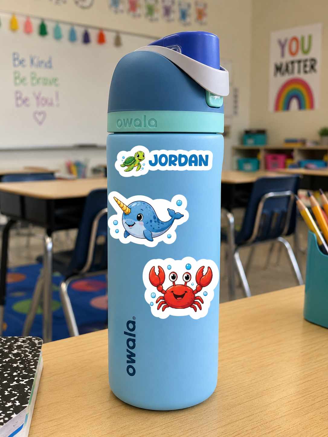 A light blue Owala water bottle with a custom name and cartoon stickers, standing on a wooden desk in a classroom.