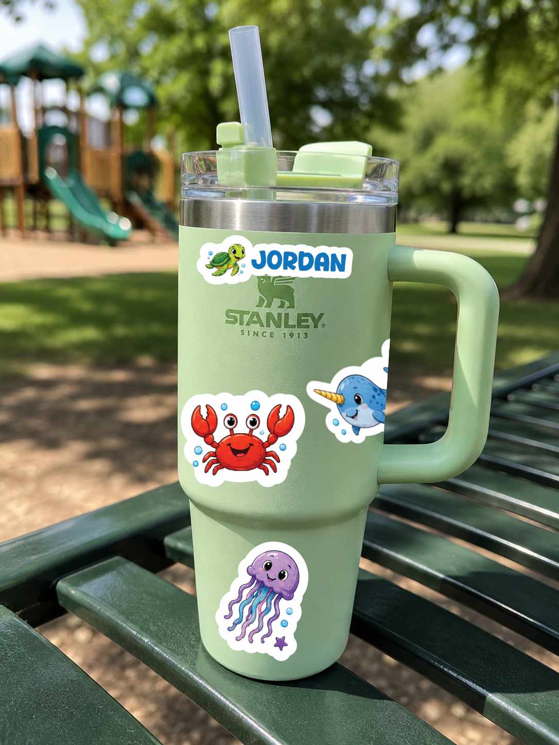 A mint green insulated travel tumbler with a handle, clear lid, and straw, placed on a green park bench outdoors. The tumbler has Jordan Stanley branding and cartoon sea-creature stickers including a red crab, blue whale, and purple octopus.