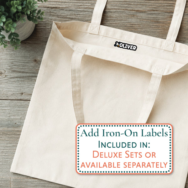 A natural canvas tote bag laid flat on a rustic wood surface, with a small black-and-white OLIVER logo patch near the top handle. A white promotional label at the bottom reads, 'Add Iron-On Labels INCLUDED IN: DELUXE SETS OR AVAILABLE SEPARATELY.