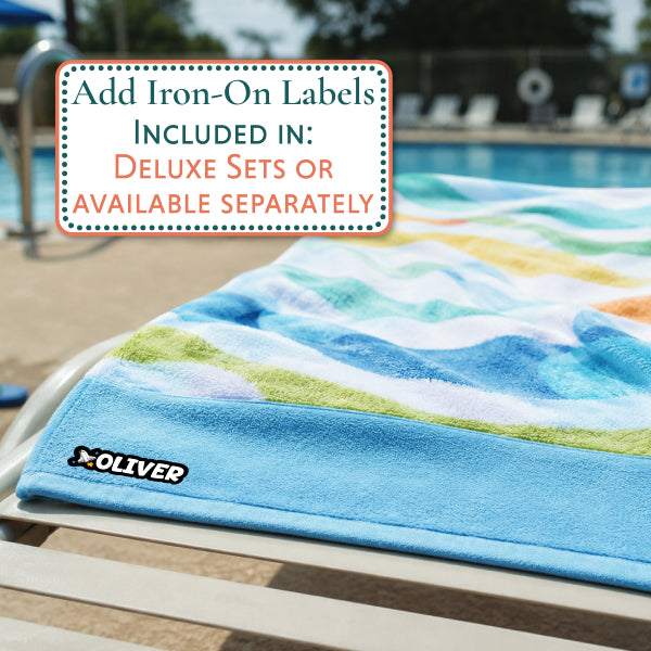 A colorful beach towel with a blue, green, and yellow landscape pattern draped over a poolside lounge chair, featuring a black OLIVER logo. A white text box in the upper left advertises iron-on labels.