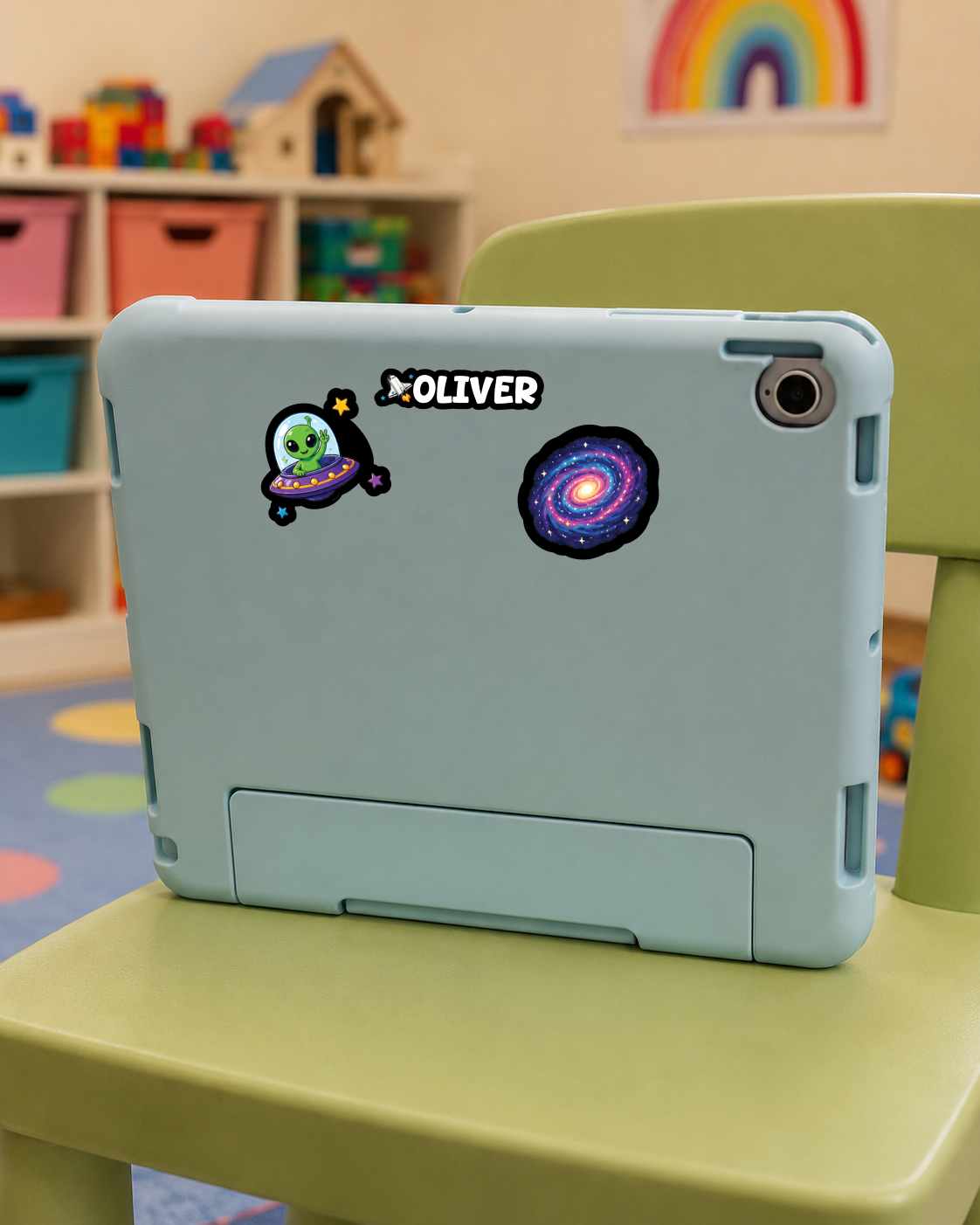 A light blue tablet case with two space-themed stickers ('Oliver' and a galaxy) on the back, resting on a green children's chair in a colorful room.