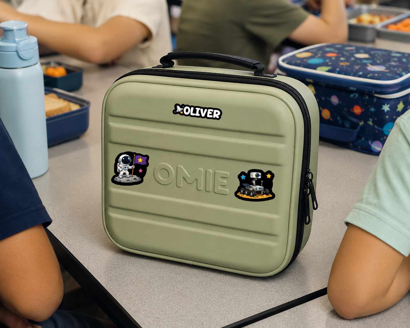 An olive green hard-shell lunch bag with a black handle and zipper, featuring the brand name OLIVER and astronaut stickers, shown on a classroom table with children seated around it.